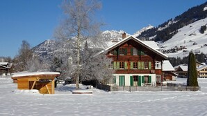 Un chalet de madera con techo inclinado, rodeado de nieve y montañas al fondo.