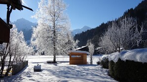 Un paisaje nevado con una cabaña de madera, un gran árbol y montañas al fondo.