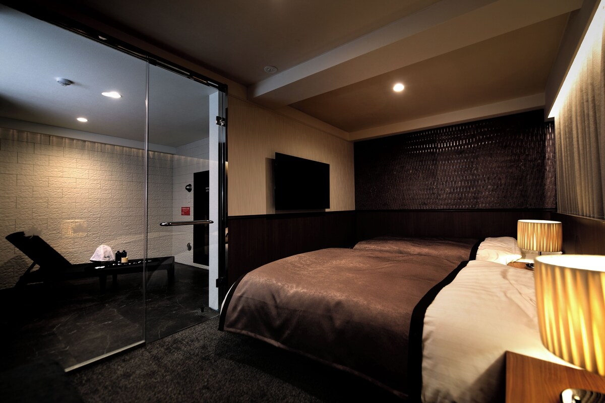 A hotel room with a bed, bedside lamps, a TV, a glass door leading to a bathroom, and a small table with toiletries.