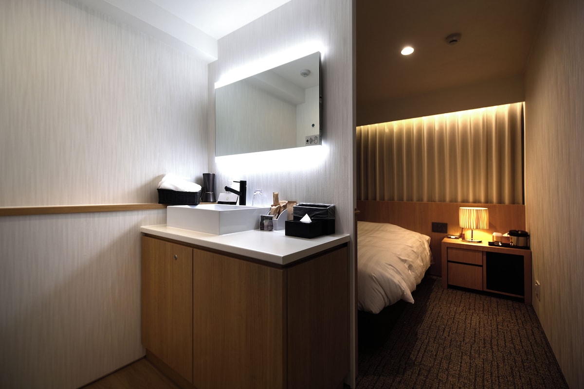 A hotel room with a bathroom featuring a sink, mirror, and cabinet. There is a bed with a headboard, a bedside table with a lamp, and a window with curtains.