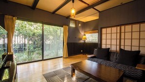 A room with a wooden floor, a sofa with cushions, a table, a lamp, and a view of greenery through large windows. - Futarishizuka (Kirishima)