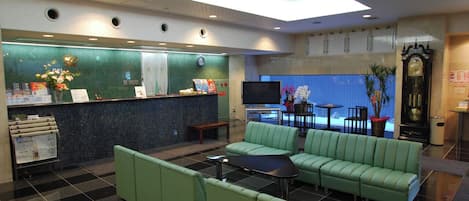 Un vestíbulo de hotel con mostrador de recepción, zona de estar verde y una pantalla de televisión.
