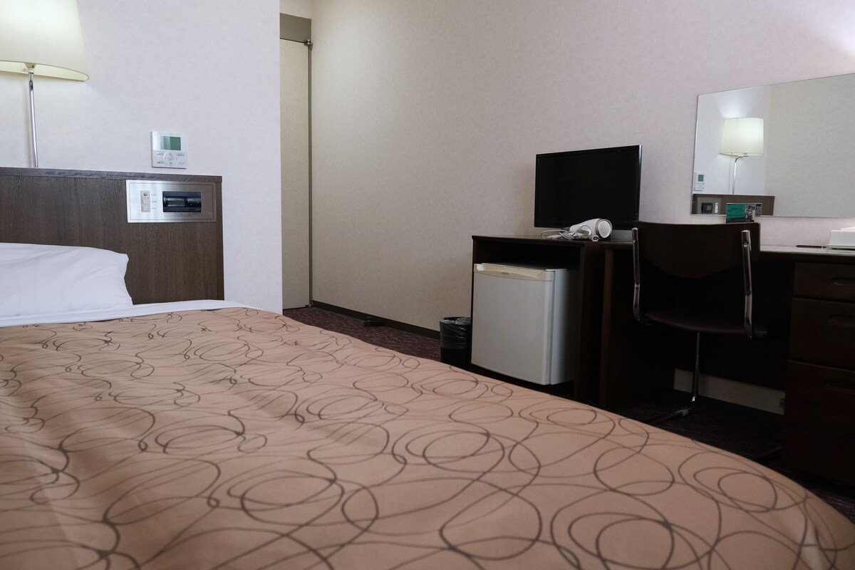 A hotel room with a bed, a desk, a chair, a small refrigerator, a TV, and a mirror.