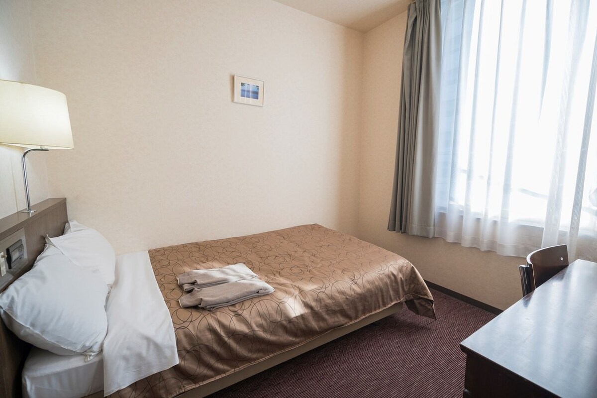 A hotel room with a bed, a desk, a lamp, and a window with curtains.
