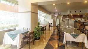 A restaurant with tables, chairs, a potted plant, and a kitchen area.