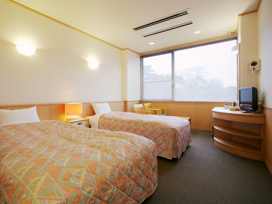 A hotel room with two beds, a desk, a chair, a TV, and a window with blinds.