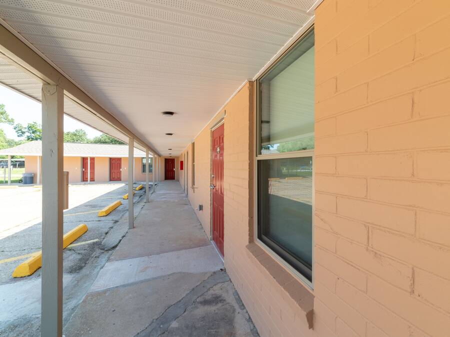A covered walkway with a brick wall and windows, leading to a building with red doors.