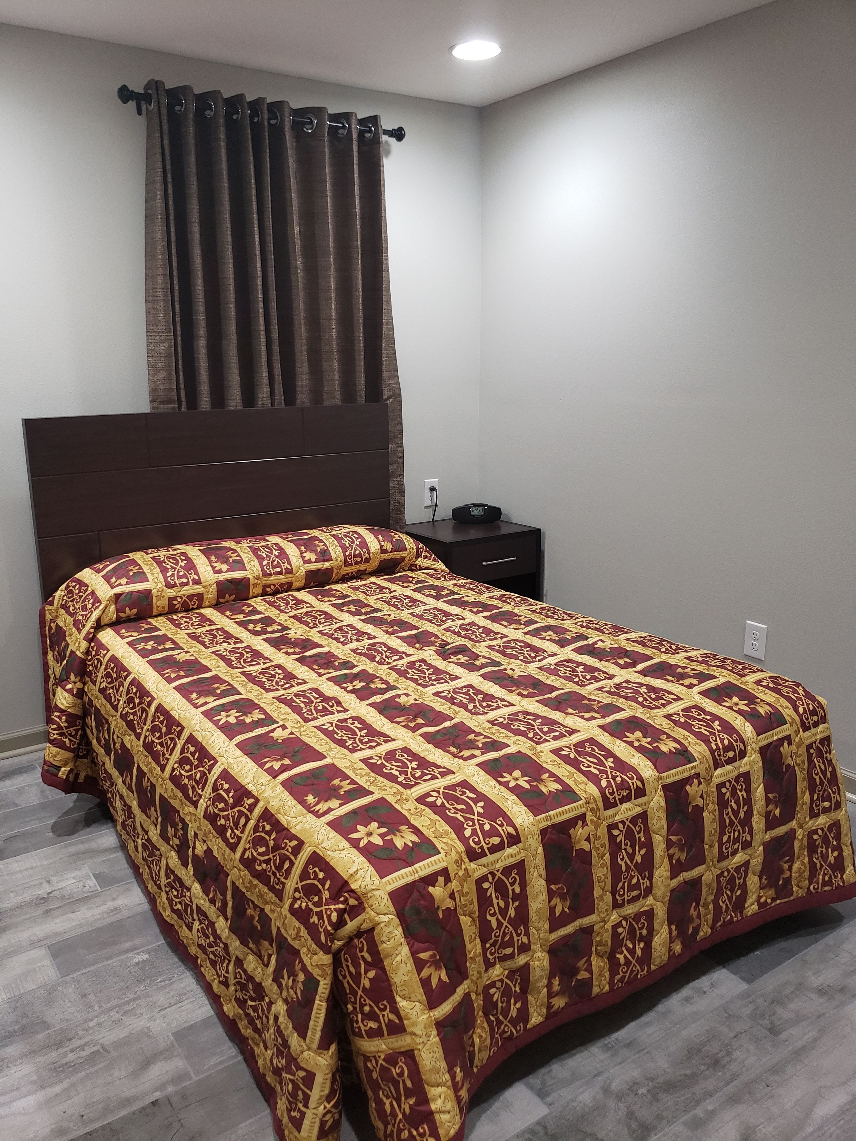 A neatly made bed with a patterned bedspread, a dark headboard, a bedside table with a radio, and a power outlet on the wall.
