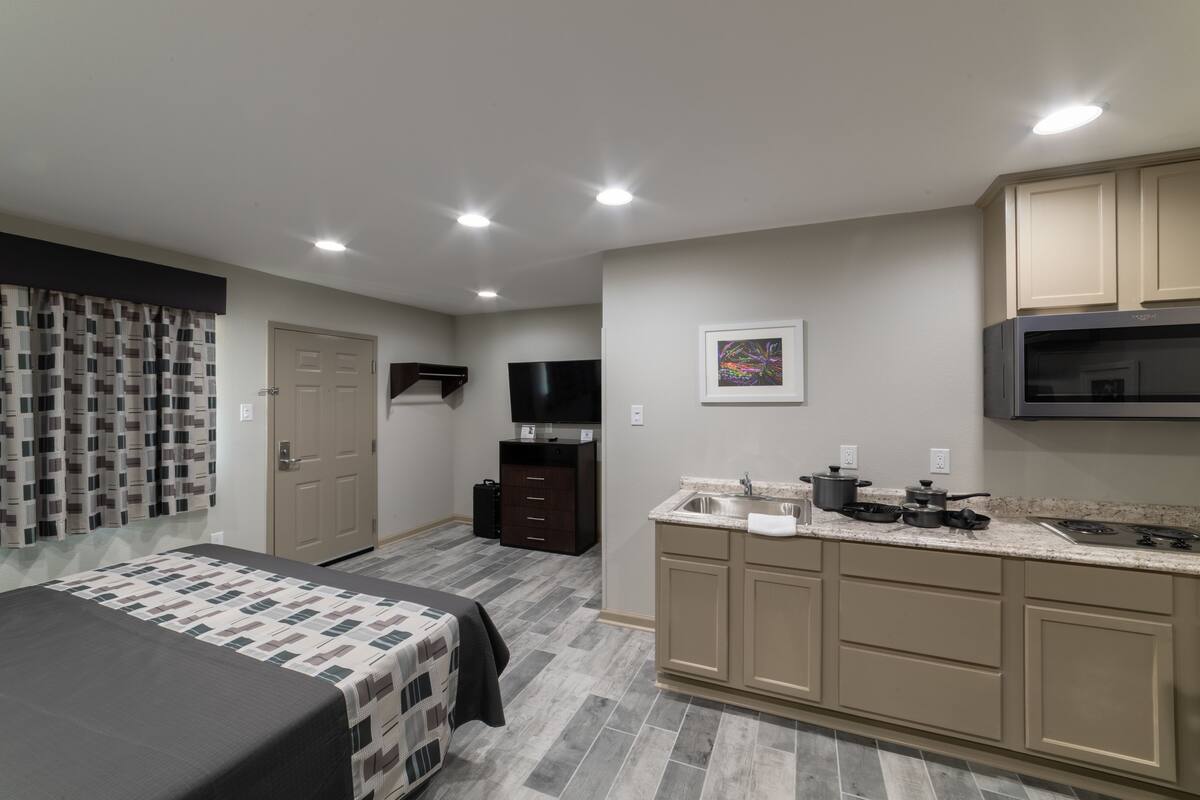 A modern hotel room with a bed, a small kitchenette, a television, and a bathroom.