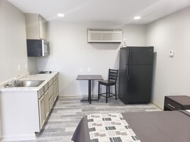 Una kitchenette compacta con lavamanos, estufa y microondas, una pequeña mesa con una silla, un refrigerador y una cama con colcha estampada.
