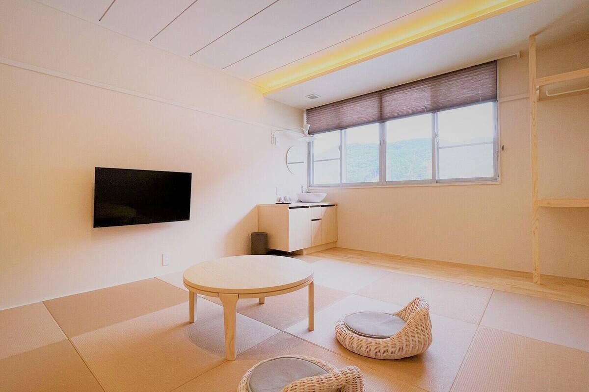 A minimalist room with a television, a small table, a wicker chair, a window with a view, and a built-in cabinet.