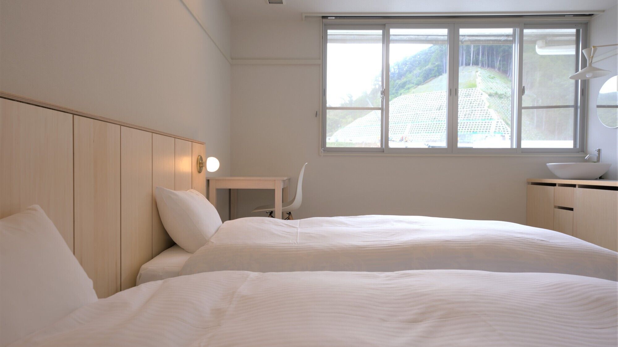 Habitación de hotel con dos camas, un escritorio, una silla, una ventana con vistas a un edificio y una lámpara de noche.