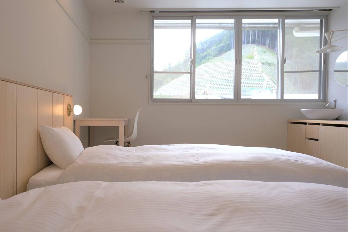 A hotel room with two beds, a desk, a chair, a window with a view of a building, and a bedside lamp.
