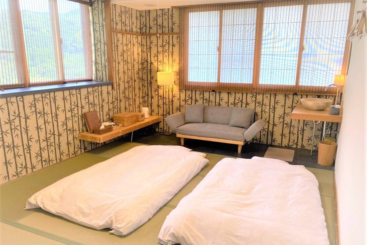 A traditional Japanese room with tatami flooring, a futon bed, a low table, a sofa, and bamboo-patterned wallpaper.