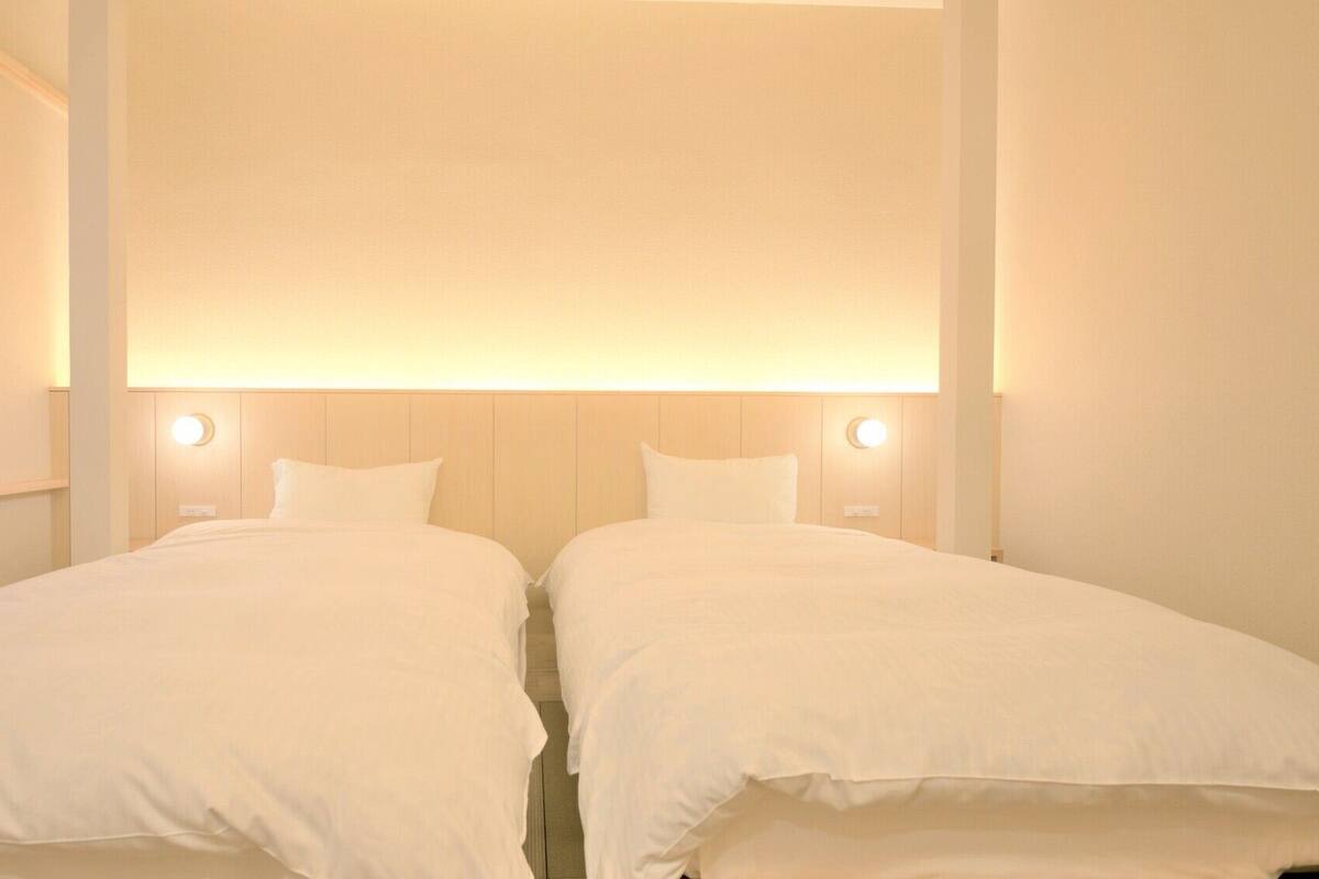 A hotel room with two beds, a large window with blinds, and wall-mounted lighting.