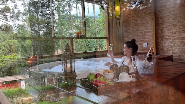 Una persona relajándose en un jacuzzi con vistas a un bosque, un muro de ladrillo y una mesa con comida y bebidas.