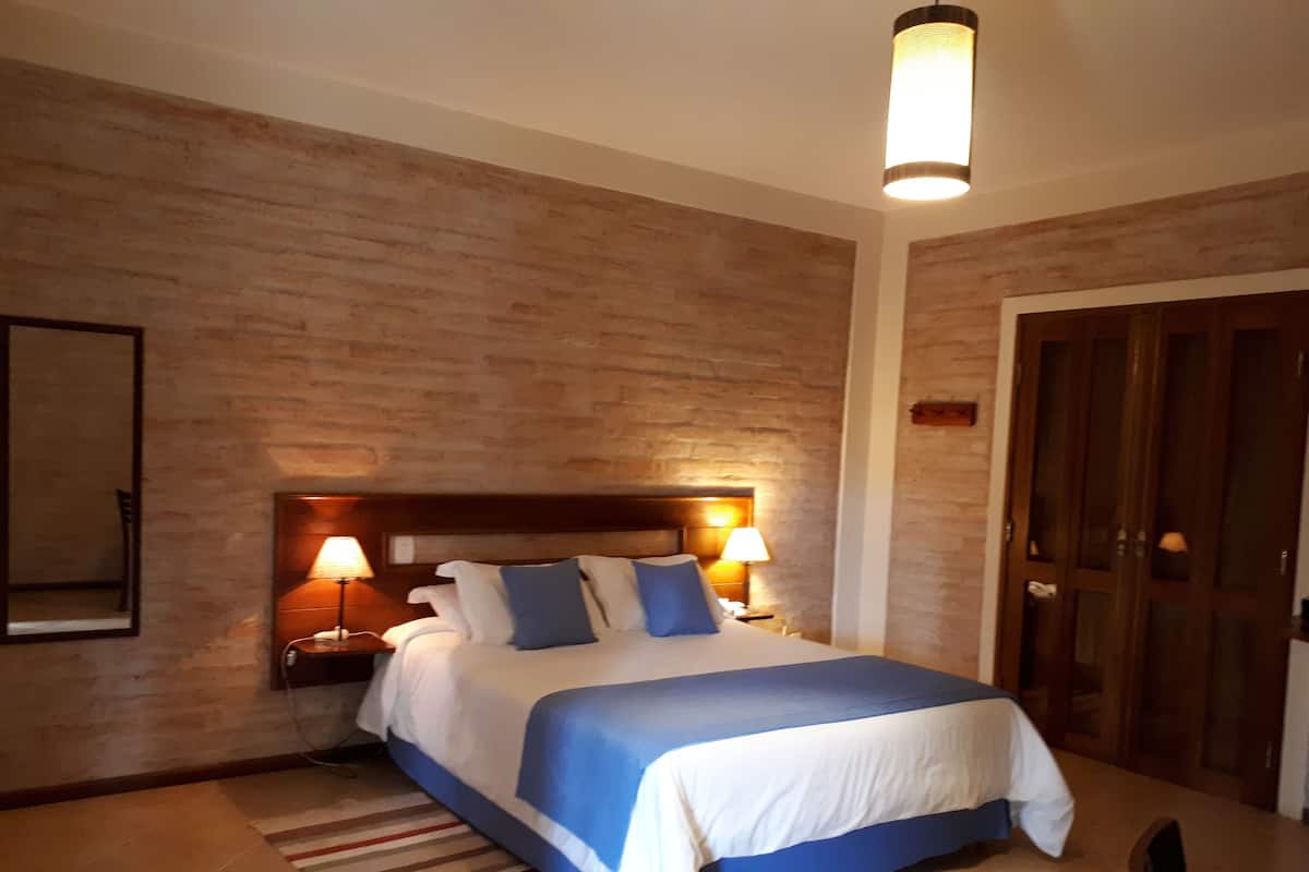 Quarto de hotel com cabeceira de madeira, cama com roupa de cama azul e branca, duas luminárias de cabeceira, um espelho e uma porta de madeira.