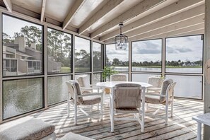 Un espace extérieur aménagé, abrité et doté de tables et de chaises, de grandes fenêtres et d’une vue sur une maison et un lac.
