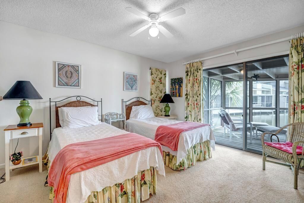 Une chambre d’hôtel avec deux lits, un ventilateur de plafond, une fenêtre avec des rideaux, une chaise et une petite table avec une lampe.