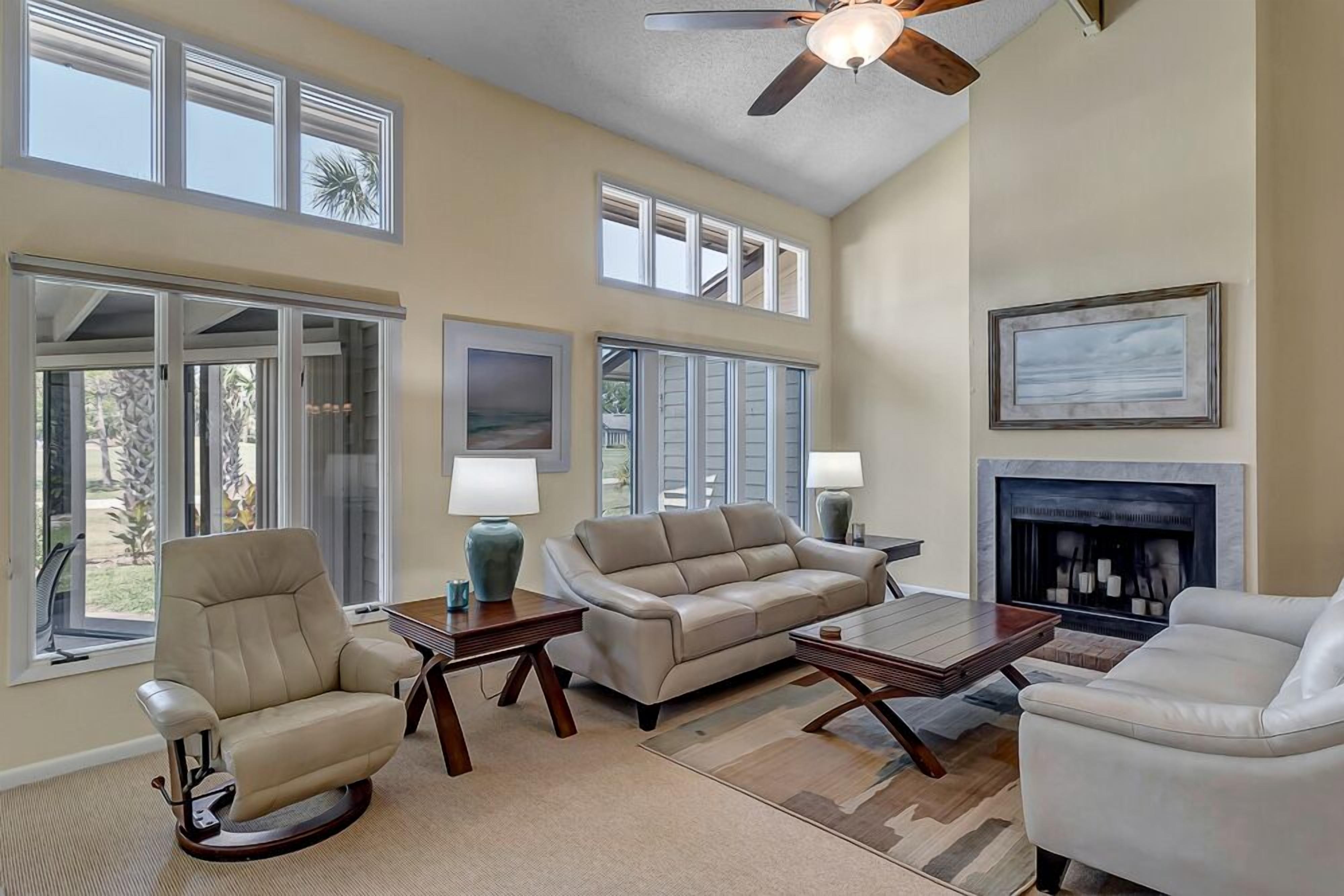 A spacious living room with a high ceiling, large windows, a fireplace, and a ceiling fan.