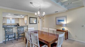 A dining area with a wooden table and white chairs, a chandelier, and a kitchen in the background. - Belle Vue - Modern condo near beach w/ golf views (Ponte Vedra Beach)