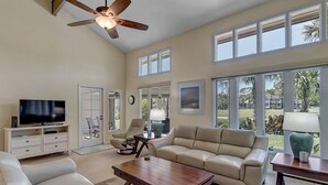 A spacious living room with a ceiling fan, a television on a stand, a beige sofa, a coffee table, and large windows offering a view of the outdoors. - Belle Vue - Modern condo near beach w/ golf views (Ponte Vedra Beach)