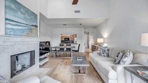 A modern living room with a fireplace, a dining area, and a kitchen in the background. - Seas the Day - Serene condo w/ pool & beach access (Ponte Vedra Beach)