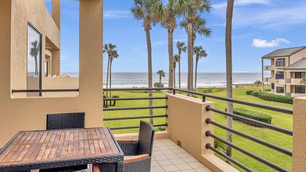 A balcony with a view of the ocean, palm trees, and a building in the background. - Seas the Day - Serene condo w/ pool & beach access (Ponte Vedra Beach)