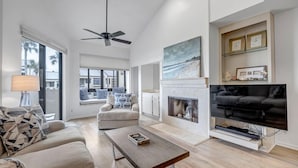 A spacious living room with a fireplace, a large TV, and a ceiling fan. - Seas the Day - Serene condo w/ pool & beach access (Ponte Vedra Beach)