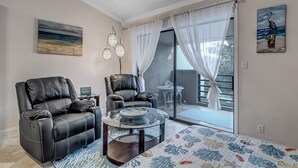 A living room with two black leather recliners, a glass coffee table, a patterned rug, a painting of a bird, and a view through a glass door to an outdoor area. - Fun in the Sun - Coastal studio w/ pool access (Ponte Vedra Beach)