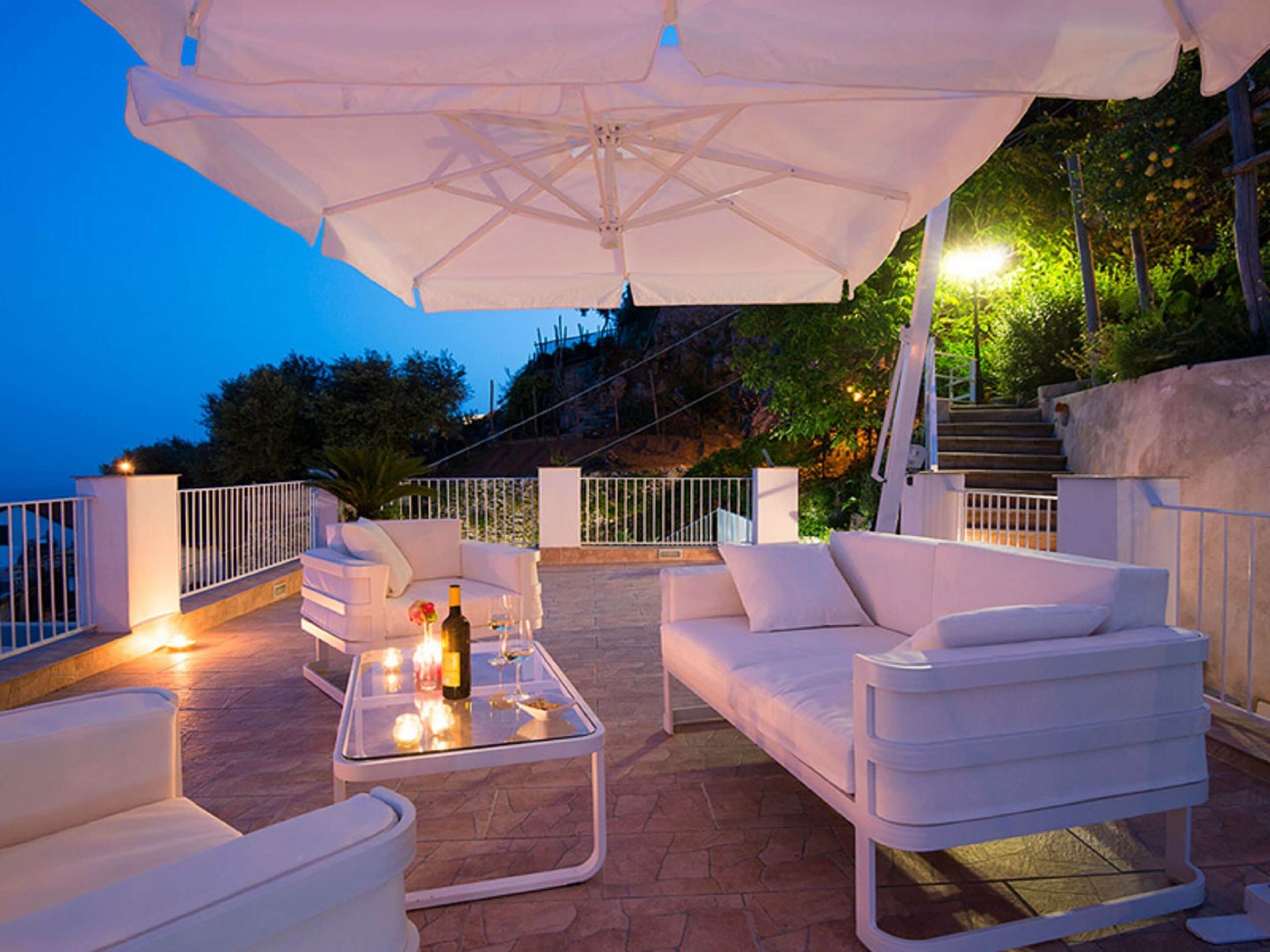 An outdoor seating area with white furniture and a glass table, under a large umbrella, overlooking a scenic hillside.