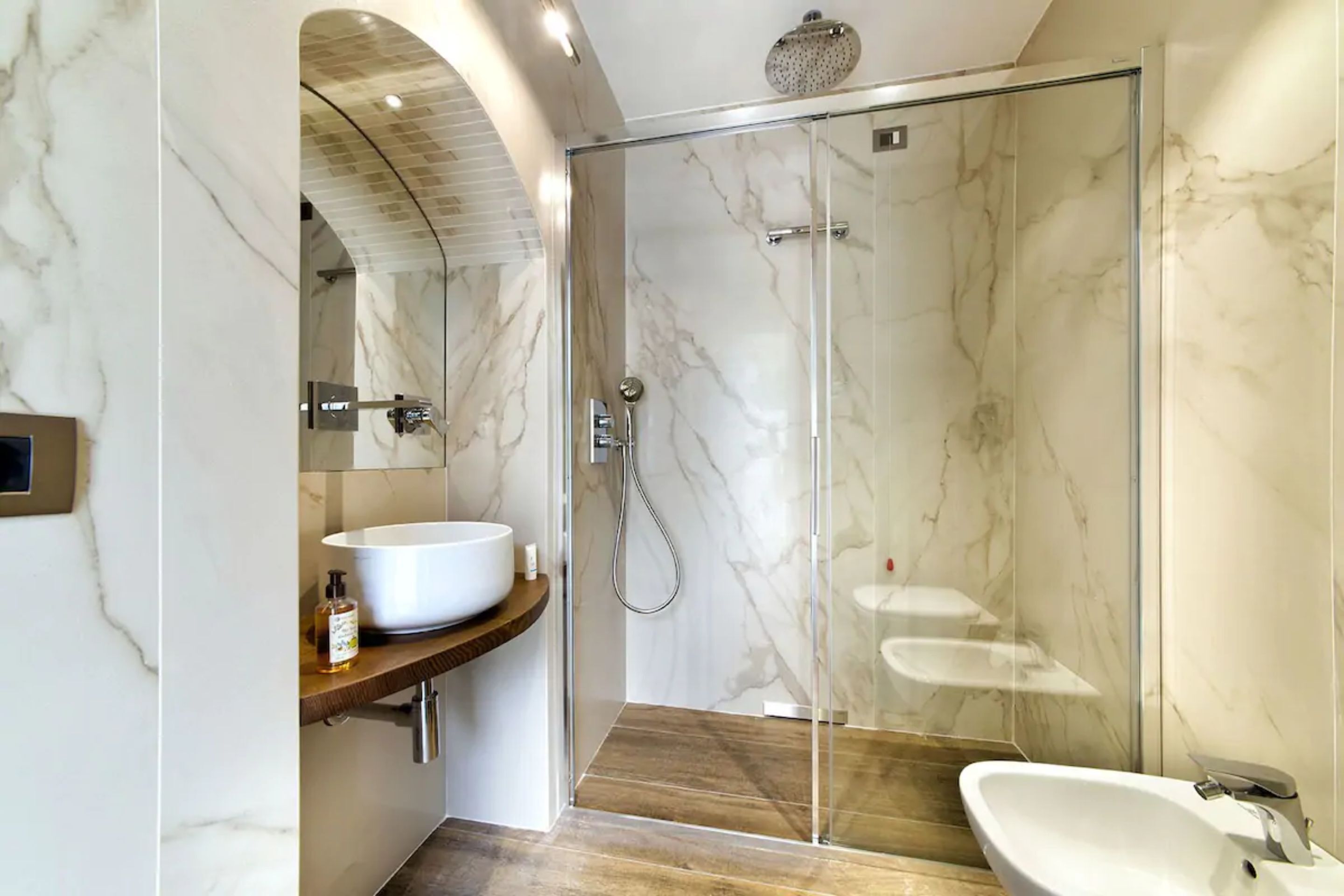 A modern bathroom with a glass-enclosed shower, a wooden vanity with a white sink, and marble walls.