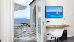 A balcony with a view of the sea, a white chair, a small table with a vase, and a mounted TV displaying a sailboat. - Villa Orleans (Amalfi)
