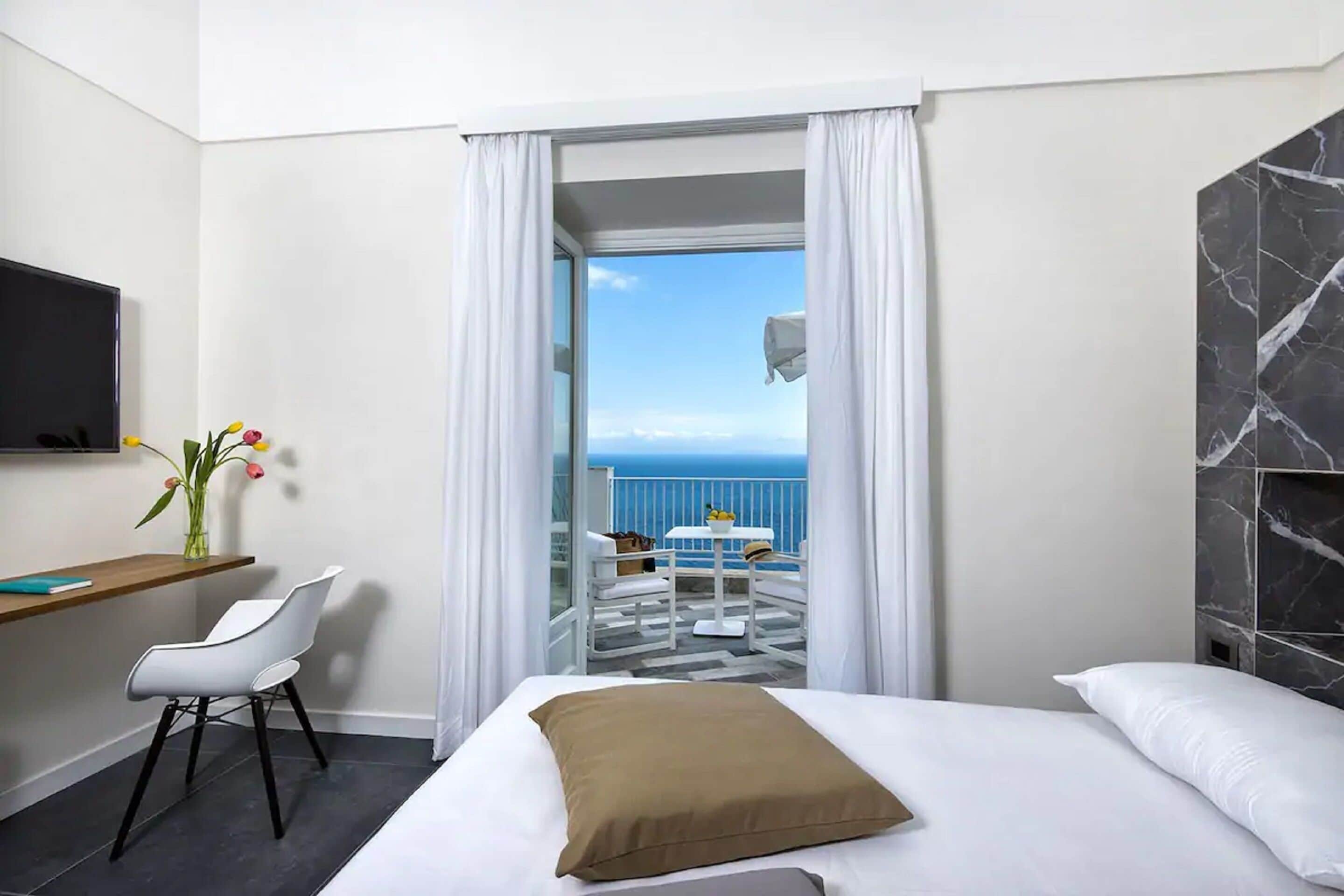 A hotel room with a bed, a desk with a chair, a TV, a vase with flowers, and a view of the sea through a glass door.