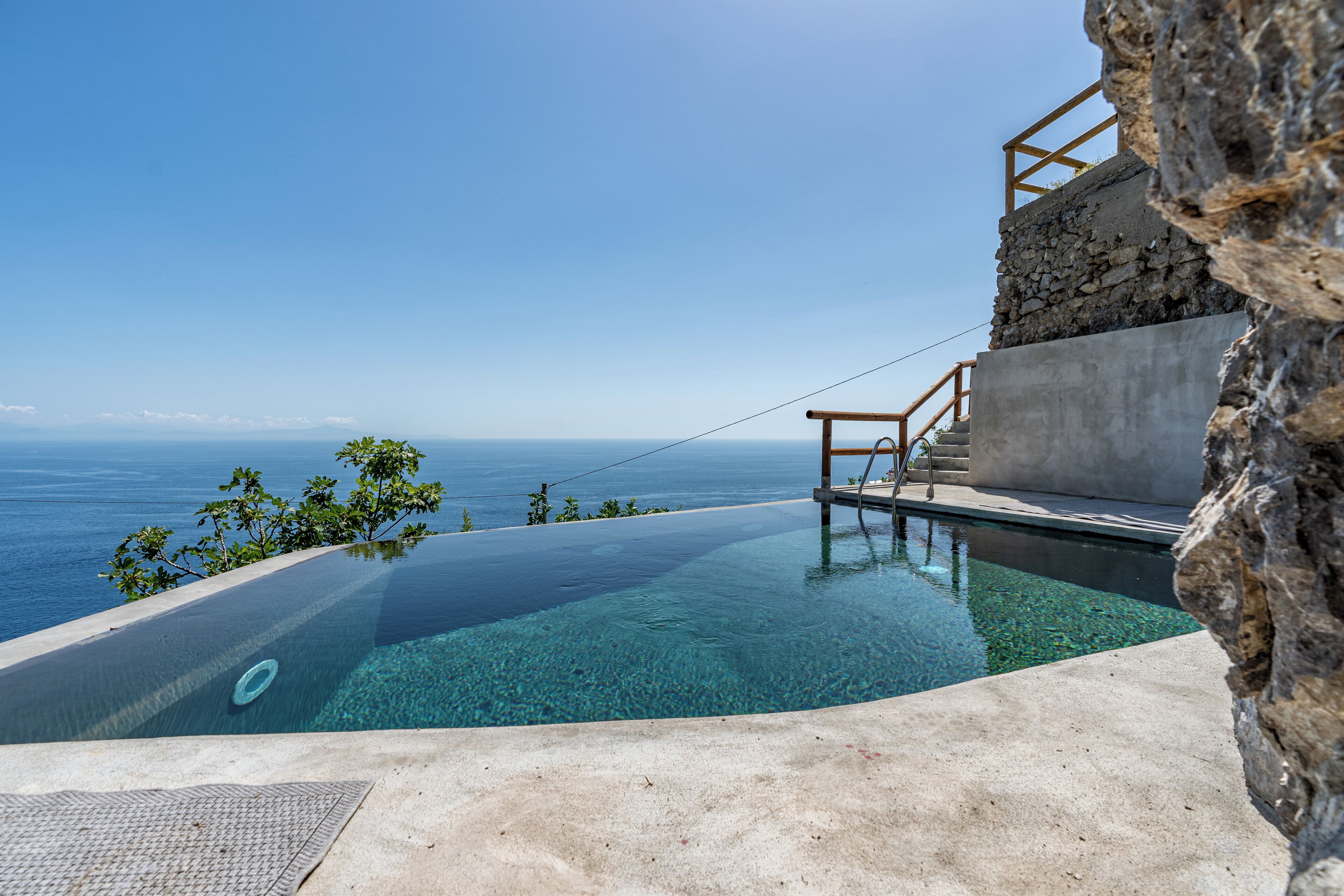 An infinity pool with a clear view of the ocean, a stone wall, and a wooden staircase.