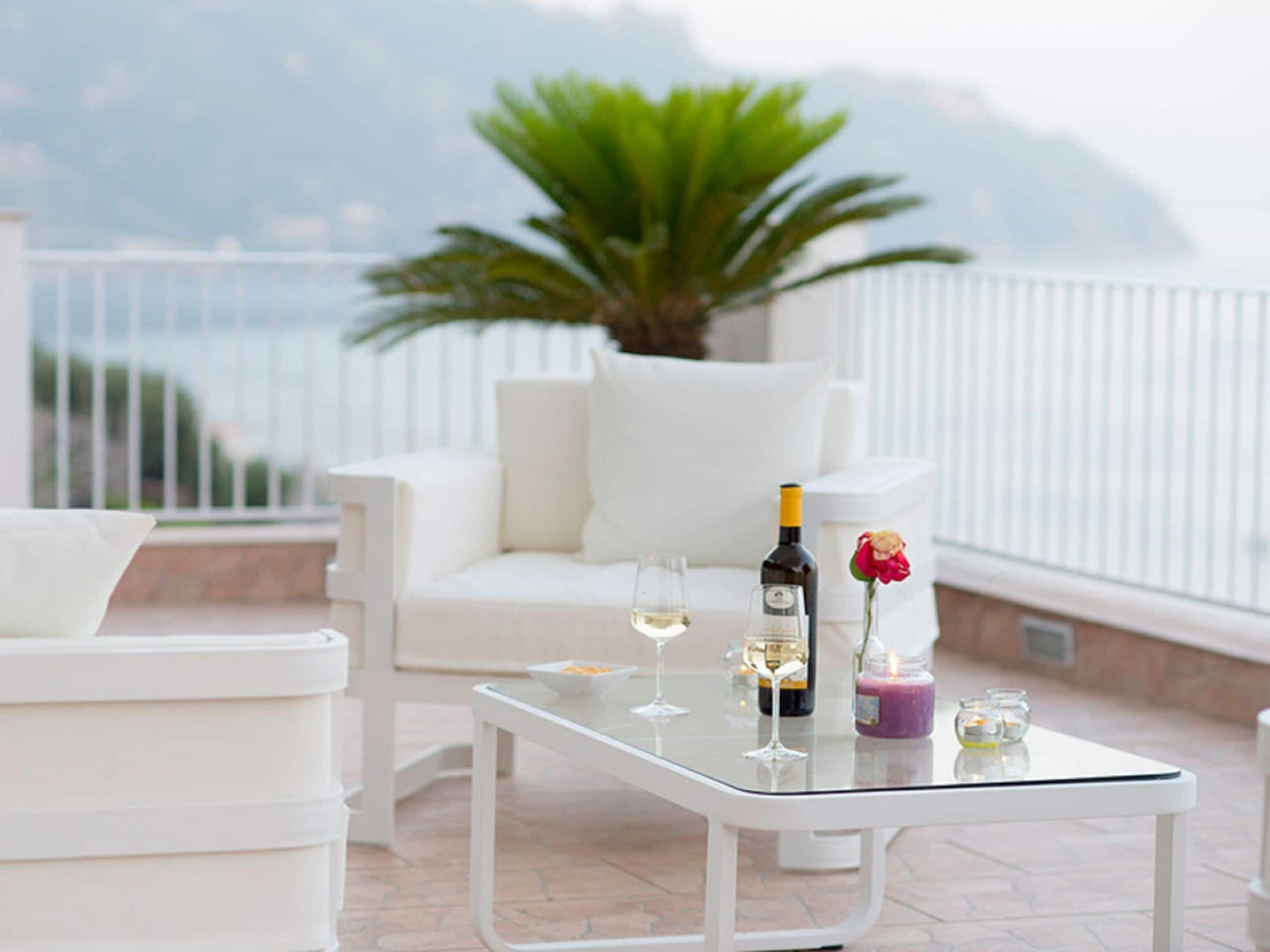 Zona salotto esterna con mobili bianchi, un tavolo in vetro, cuscini bianchi, una pianta in vaso e una bottiglia di vino.