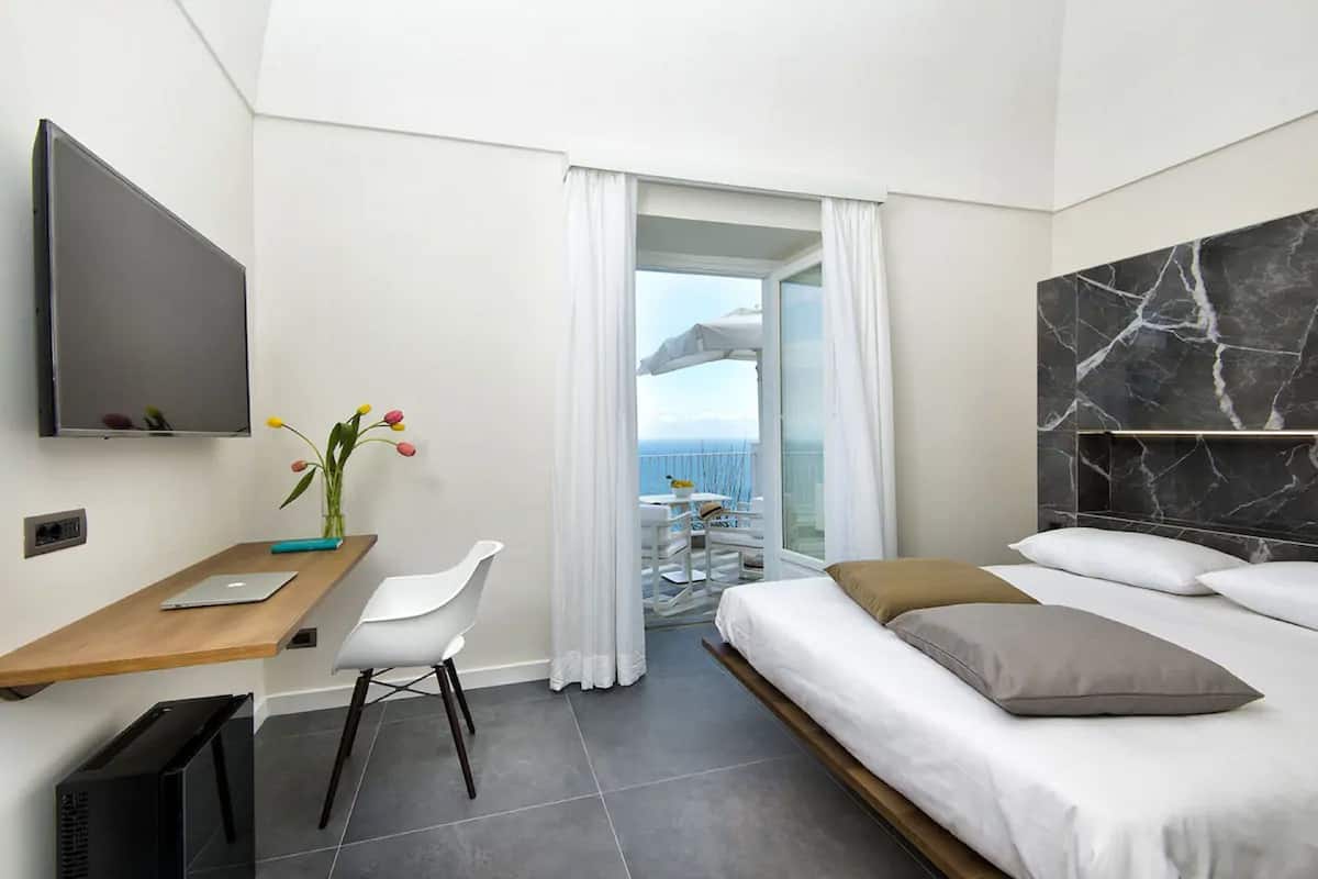 Una moderna camera d'albergo con un letto, una scrivania con un portatile, una televisione e un balcone con vista sul mare.
