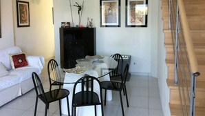 Una sala de estar moderna con un sofá blanco, una mesa de comedor de vidrio y sillas negras. Se ve una escalera a la derecha y cuadros en la pared.