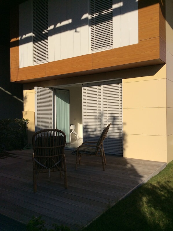 A modern building with a wooden deck, two wicker chairs, and a door leading to an interior space.