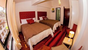 Une chambre d’hôtel avec deux lits, un plancher en bois, une tête de lit rouge, une petite table avec une lampe et une fenêtre avec des rideaux.