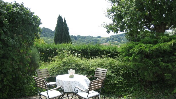Ein runder Tisch mit weißer Tischdecke und vier Stühlen, umgeben von einer Hecke und einer Pflasterterrasse.