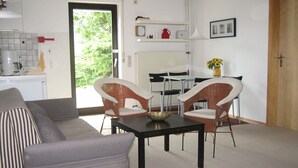 Ein Wohnzimmer mit einer Couch, zwei Korbstühlen, einem Couchtisch und einem Esstisch mit Stühlen. Es gibt eine Küchenzeile mit einem Kühlschrank, einer Kaffeemaschine und einem Waschbecken. Eine Tür führt in einen Außenbereich mit sichtlicher Bepflanzung.