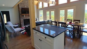 Une cuisine spacieuse dotée d’une cheminée en pierre, de poutres apparentes et de grandes fenêtres offrant une vue sur l’extérieur.