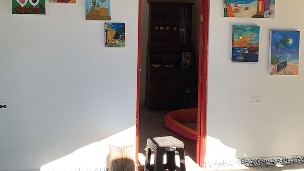 A room with a wooden chair, a small stool, and a cabinet with dishes. There are several paintings on the wall, and a door leading to another room. - Casa Campomar (Juan de Acosta)
