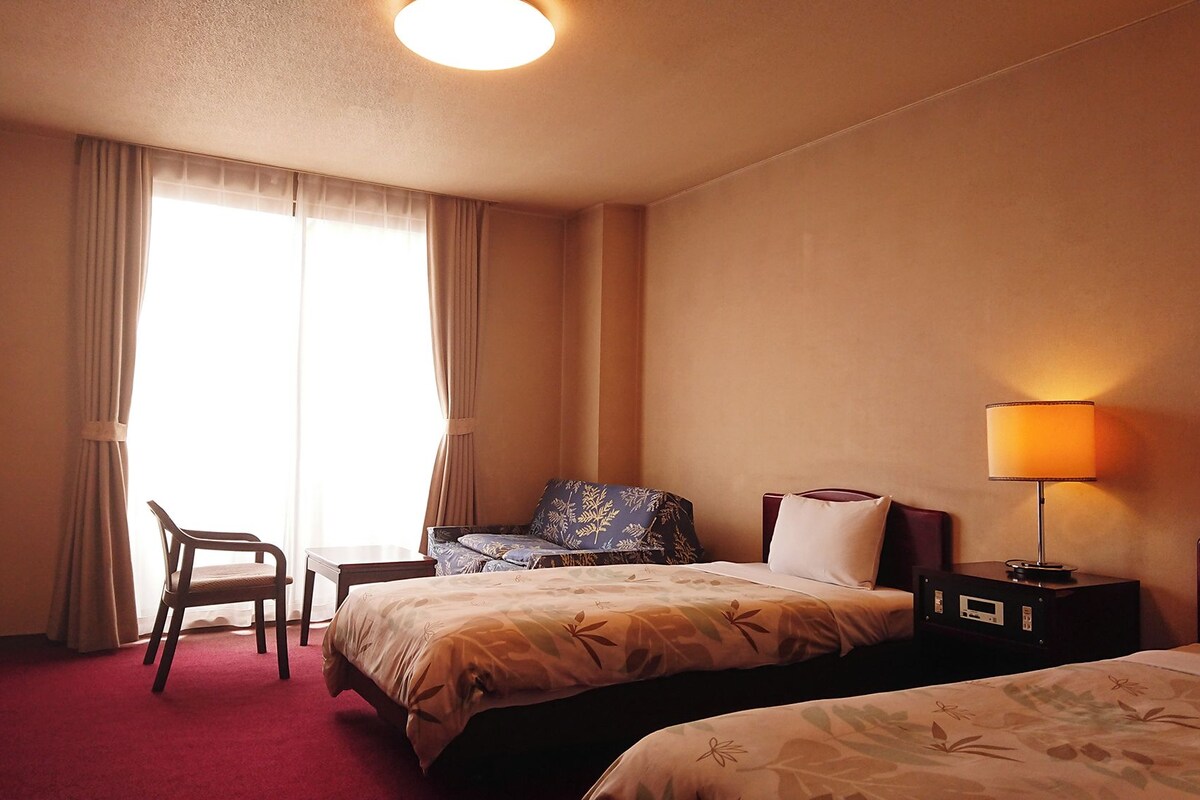 A hotel room with two beds, a chair, a desk, and a lamp.
