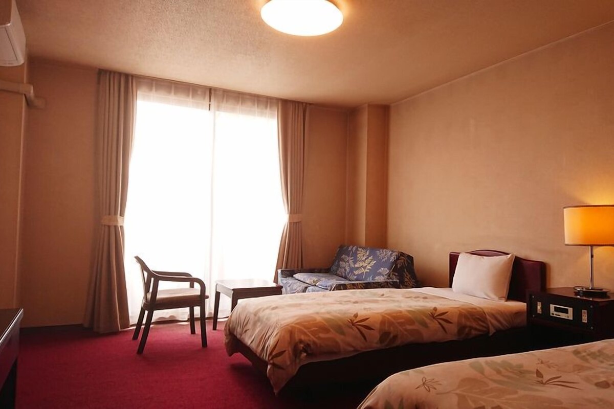 A hotel room with two beds, a desk, a chair, and a lamp.