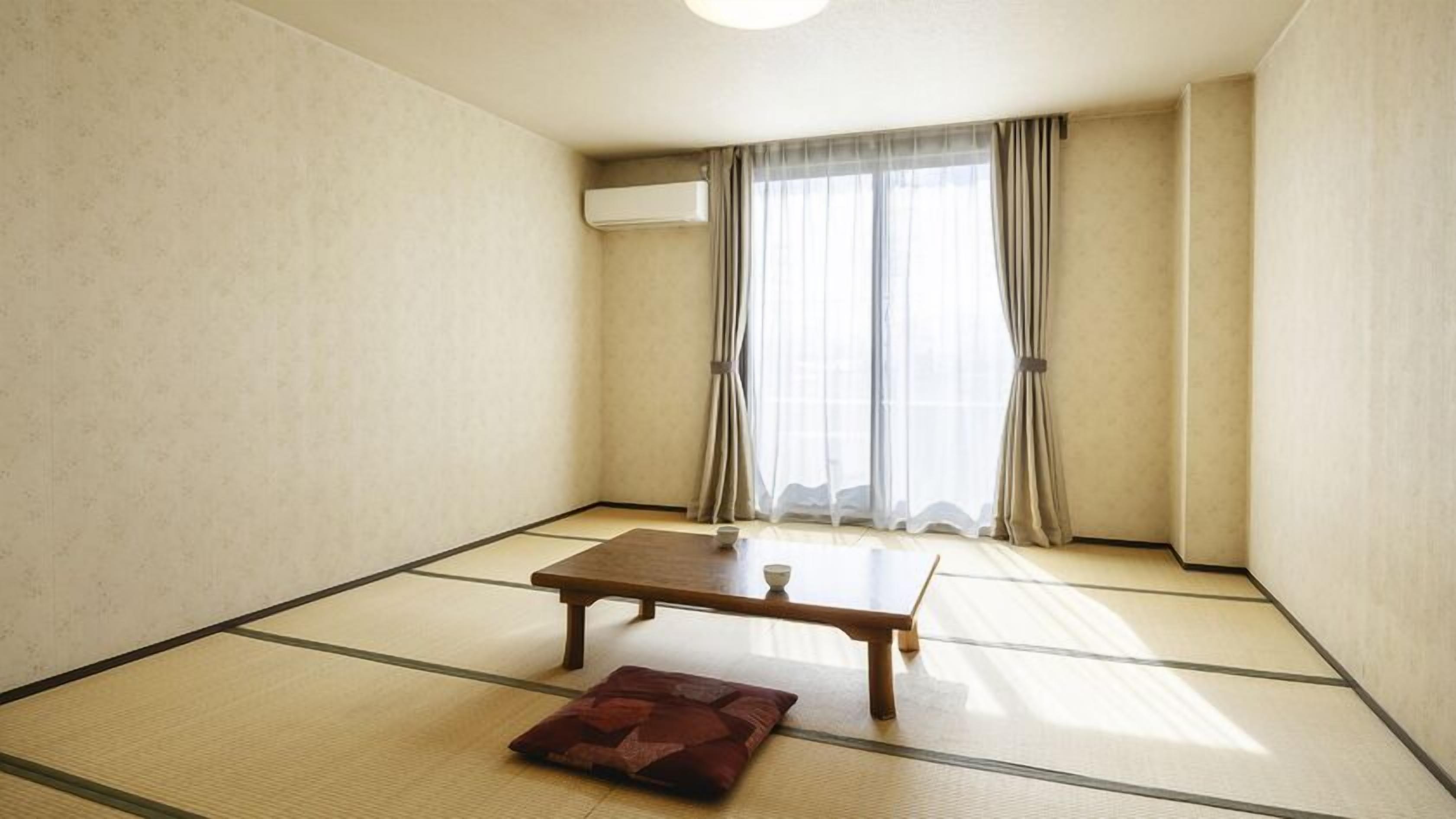 Una habitación con suelo de tatami, una mesa de madera, un cojín y una ventana con cortinas.