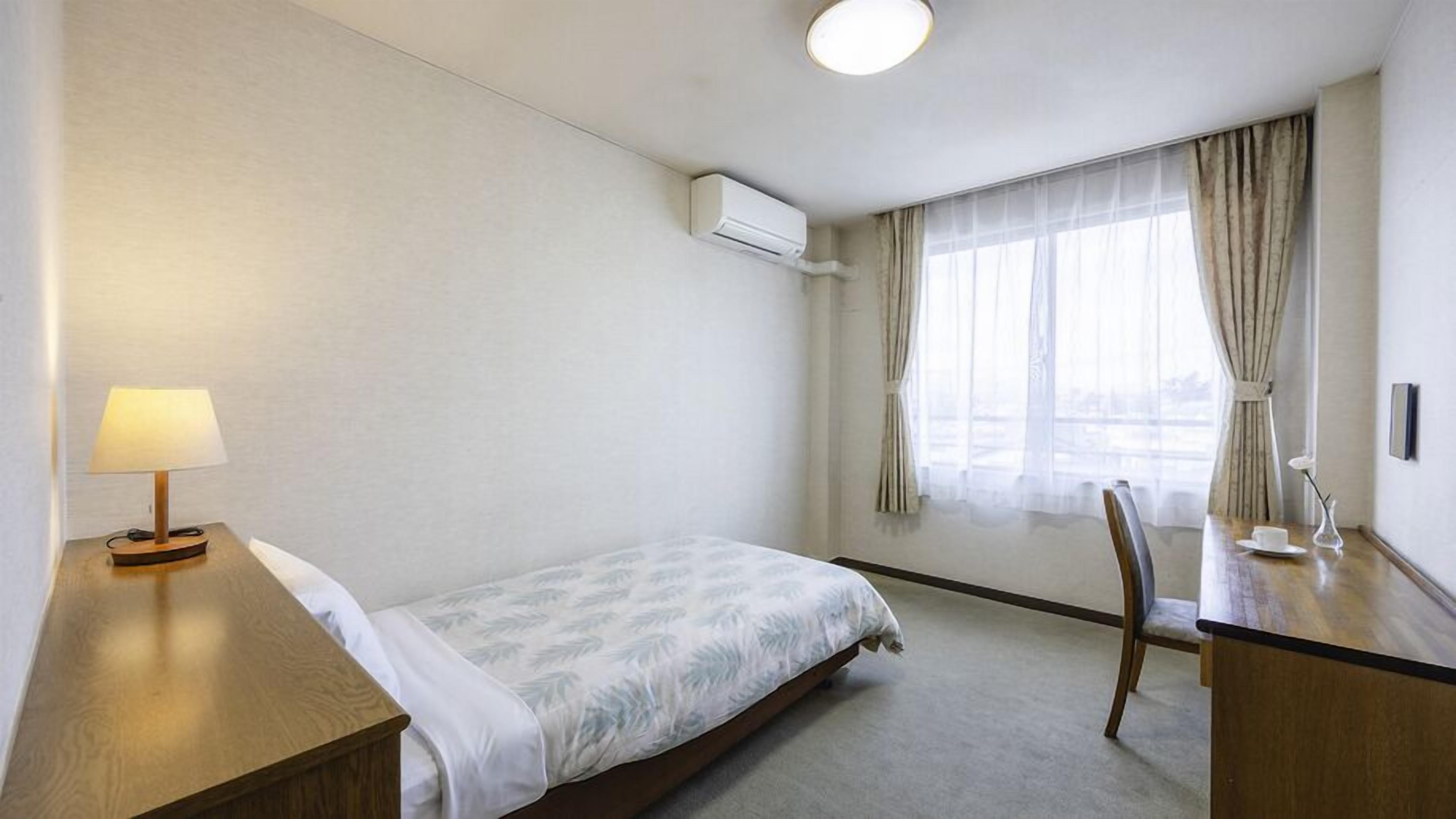 Habitación de hotel con cama, escritorio con lámpara, silla, ventana con cortinas y unidad de aire acondicionado.