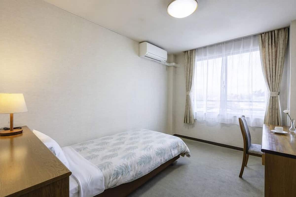 A hotel room with a bed, a desk with a lamp, a chair, a window with curtains, and an air conditioning unit.