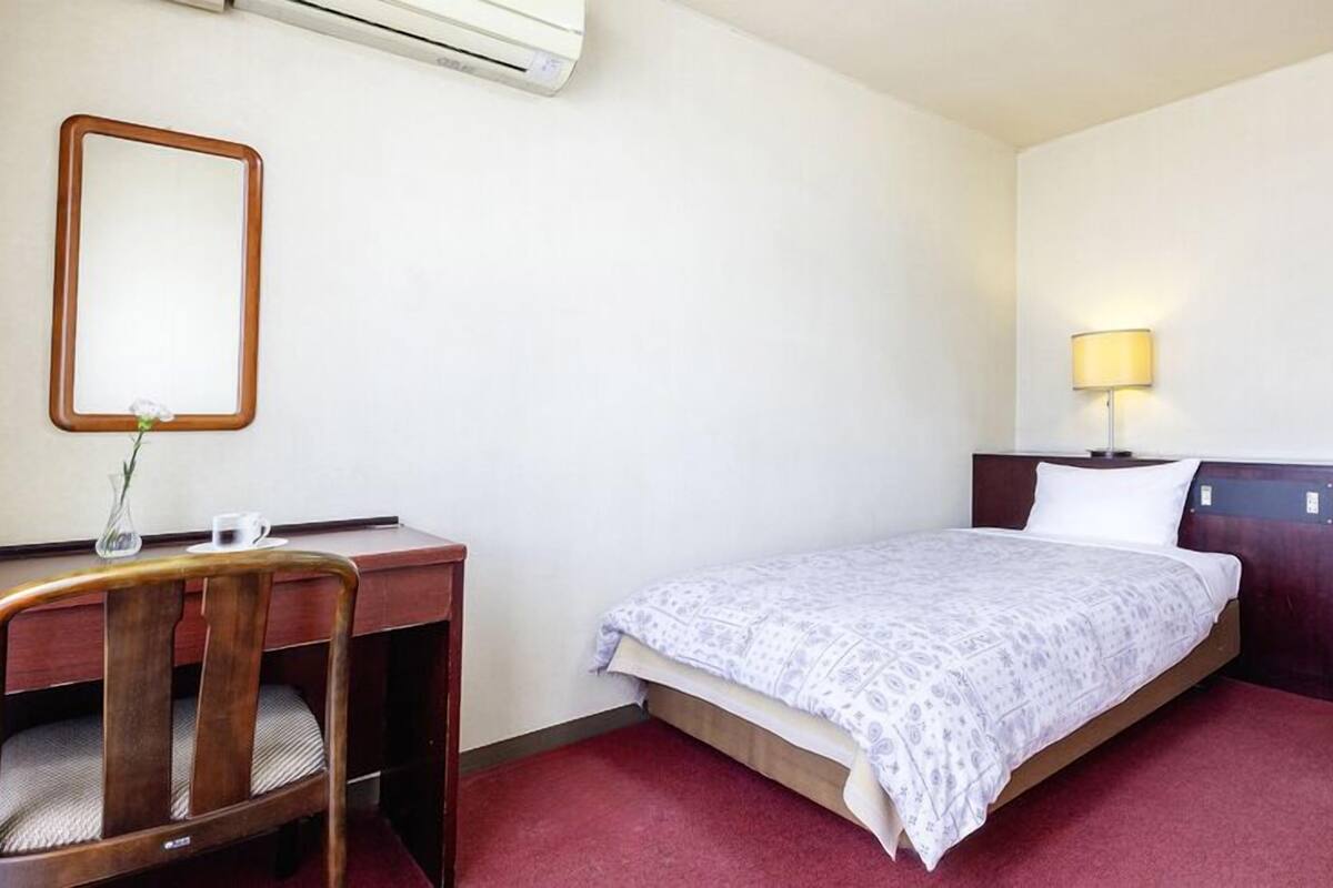 A hotel room with a bed, a desk, a chair, a lamp, and an air conditioning unit.